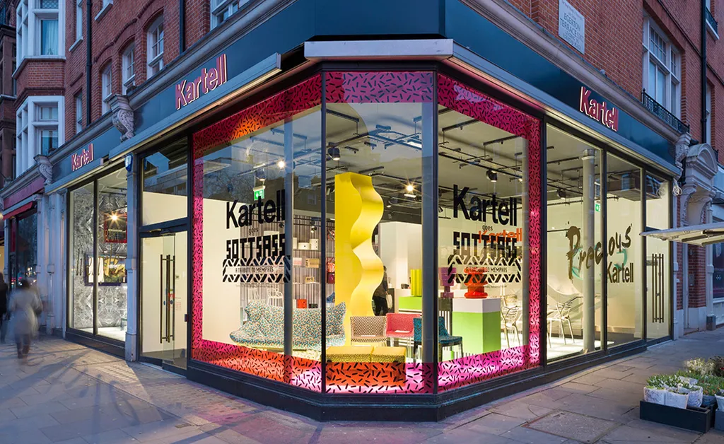 Kartell retail storefront with illuminated window display and bold pink graphic framing at a street corner.