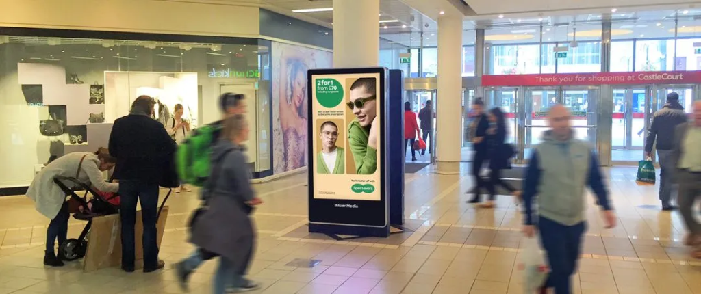 Freestanding digital advertising display inside a shopping mall with blurred shoppers walking past.