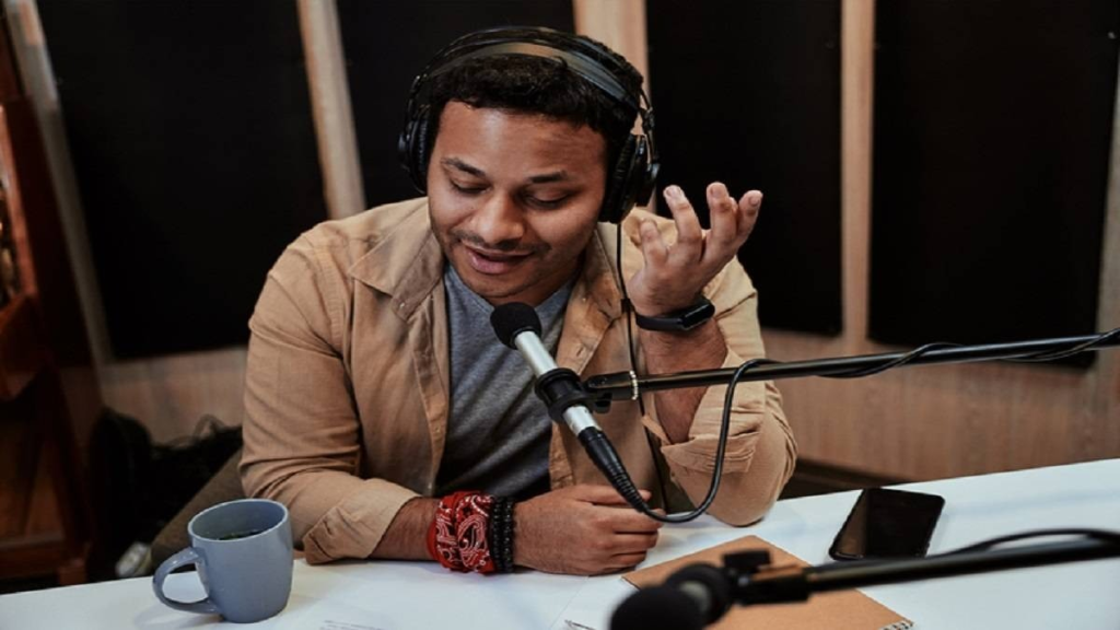 Male podcast host wearing headphones speaking into a studio microphone at a recording desk.