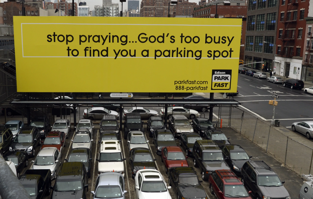 Large yellow billboard above parking lot reading “Stop praying… God’s too busy to find you a parking spot” promoting ParkFast parking service.