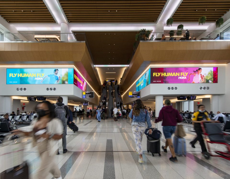 Busy airport terminal with travelers walking past large digital advertising screens displaying the “Fly Human Fly” HOKA running campaign near escalators.