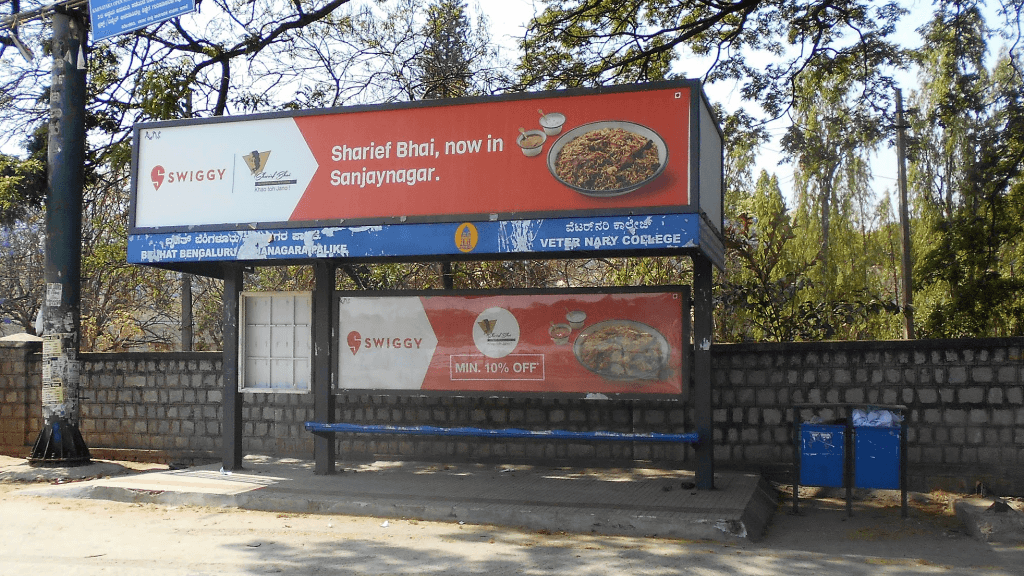 Swiggy food delivery advertisement placed at a bus shelter featuring local restaurant promotion and discount offer.
