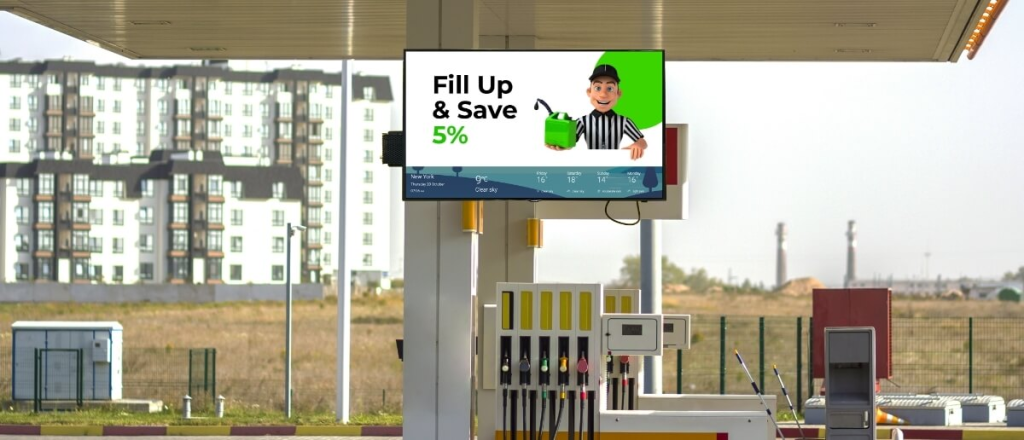 Digital advertising screen mounted above fuel pumps at a petrol station promoting a fuel discount offer while cars refuel nearby.