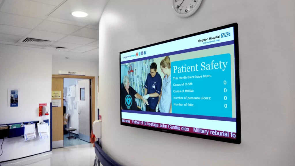 Wall-mounted digital screen in a hospital corridor displaying patient safety statistics and healthcare information.