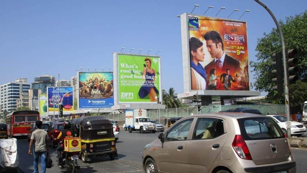 High-visibility billboards at a crowded city intersection featuring entertainment and lifestyle brands, with cars, auto-rickshaws, and pedestrians in motion.