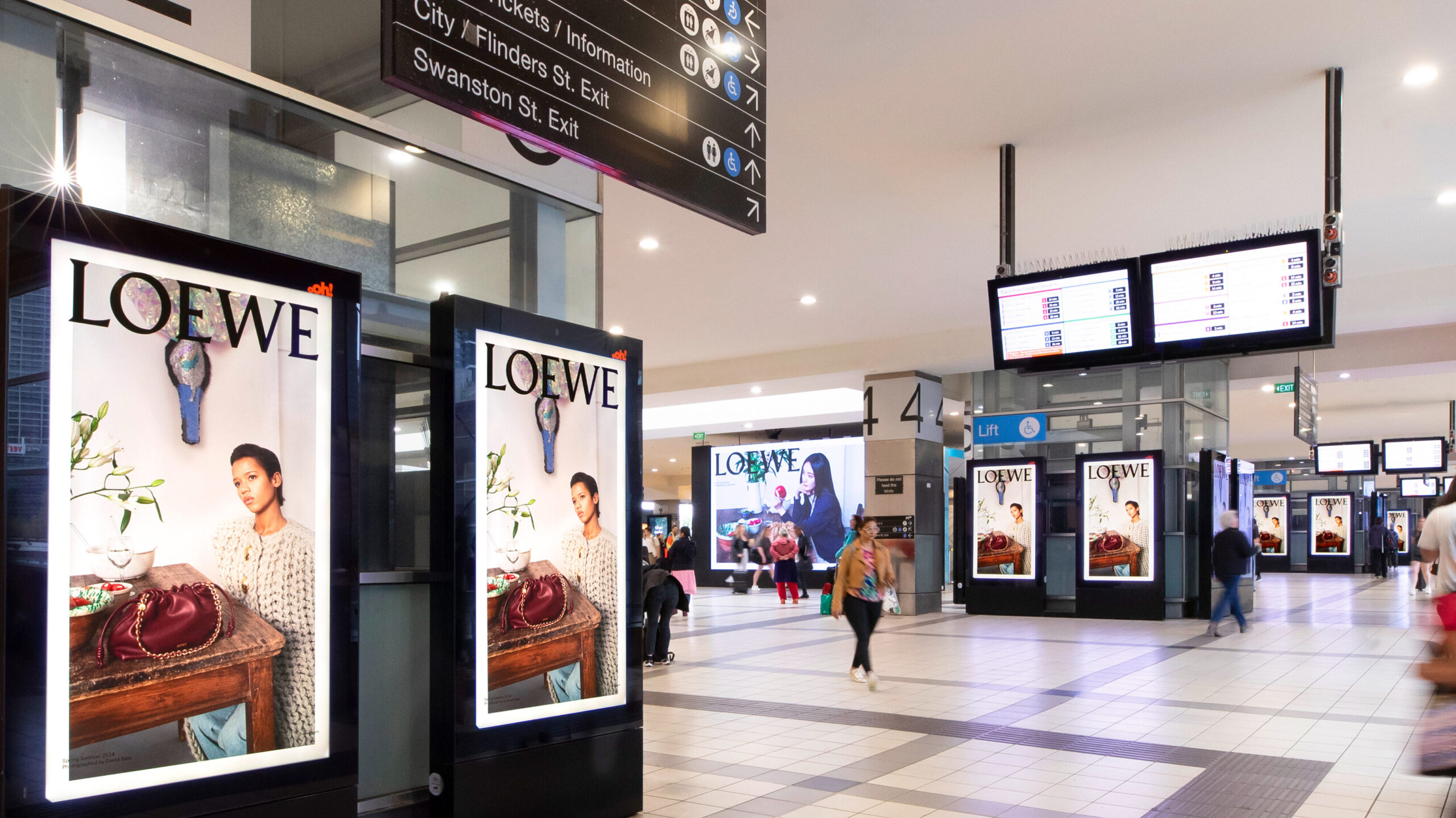 Multiple illuminated digital advertising panels featuring luxury fashion brand campaigns installed inside a busy transit station concourse.