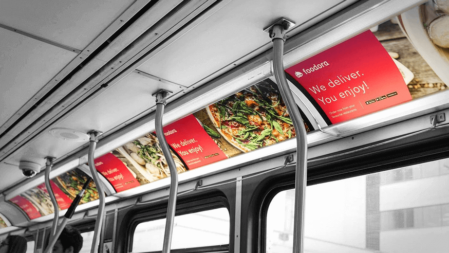 Foodora food delivery advertisement displayed inside a city bus with red panels showing dishes and the message “We deliver. You enjoy.”