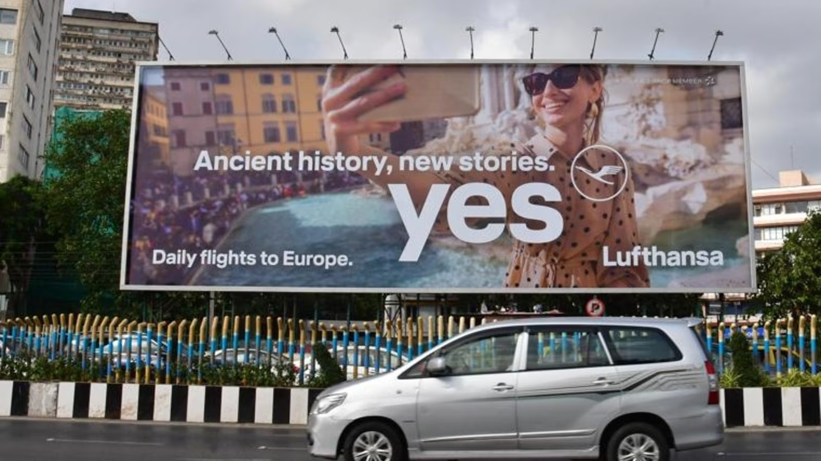 Lufthansa roadside billboard featuring a travel-themed campaign installed above a city road with moving vehicles in the foreground.