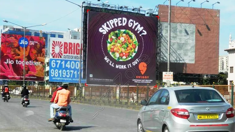 Swiggy billboard reading “Skipped Gym? We’ll Work It Out” displayed along a busy road with cars and motorcycles passing underneath.
