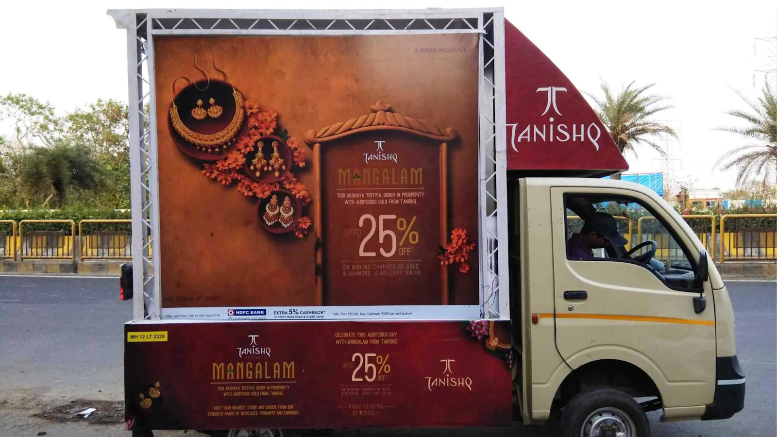 Small advertising truck carrying a large Tanishq jewellery promotional display for Mangalam offers driving on a city road.