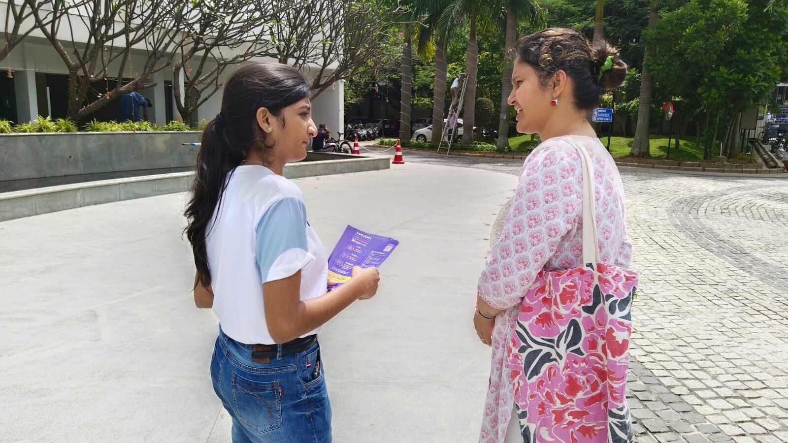 Young woman handing a promotional flyer to another woman in a residential outdoor setting as part of direct marketing activity.