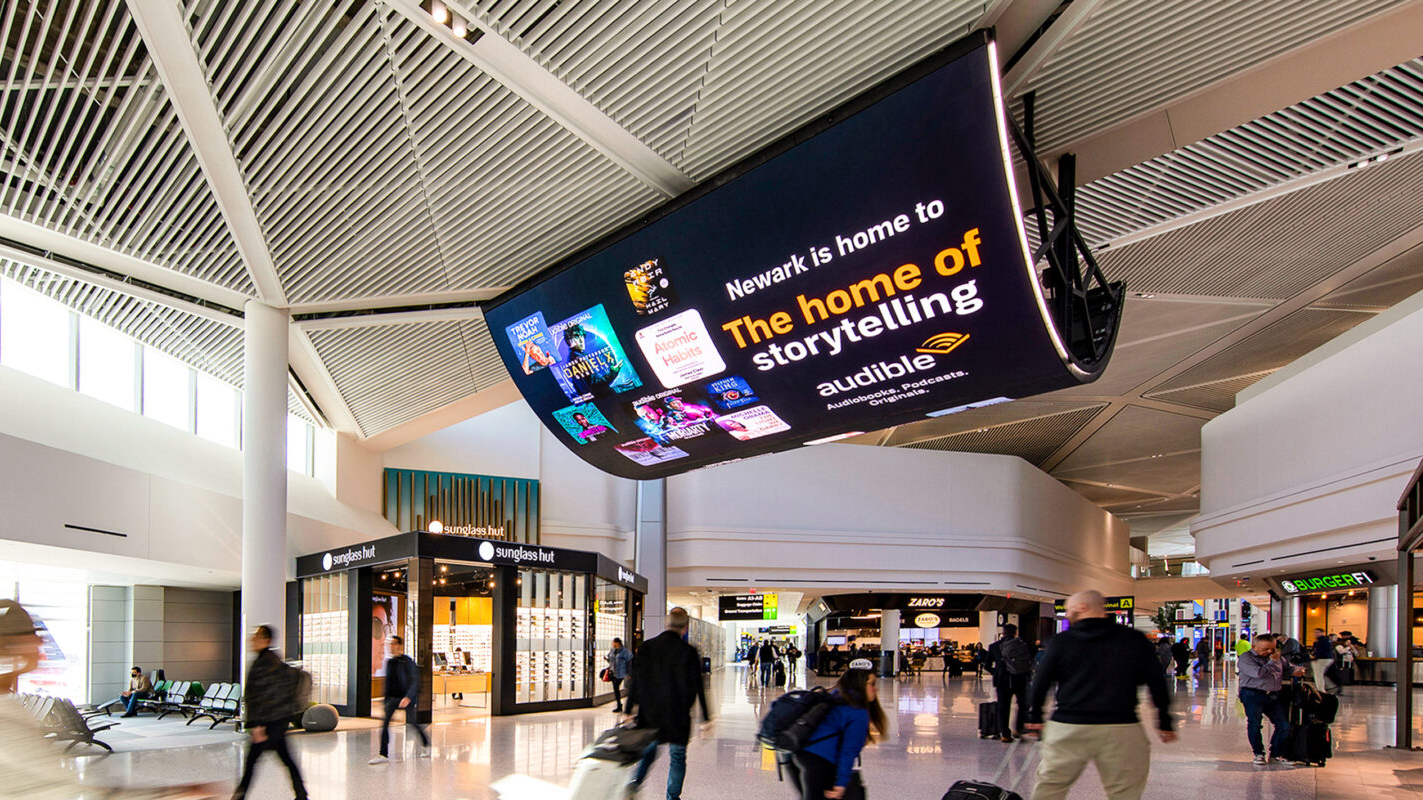 Digital advertising screen inside airport promoting Audible audiobooks with travelers walking through terminal
