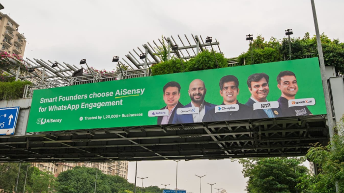 Large green bridge billboard advertising AiSensy WhatsApp engagement platform featuring founders and brand messaging